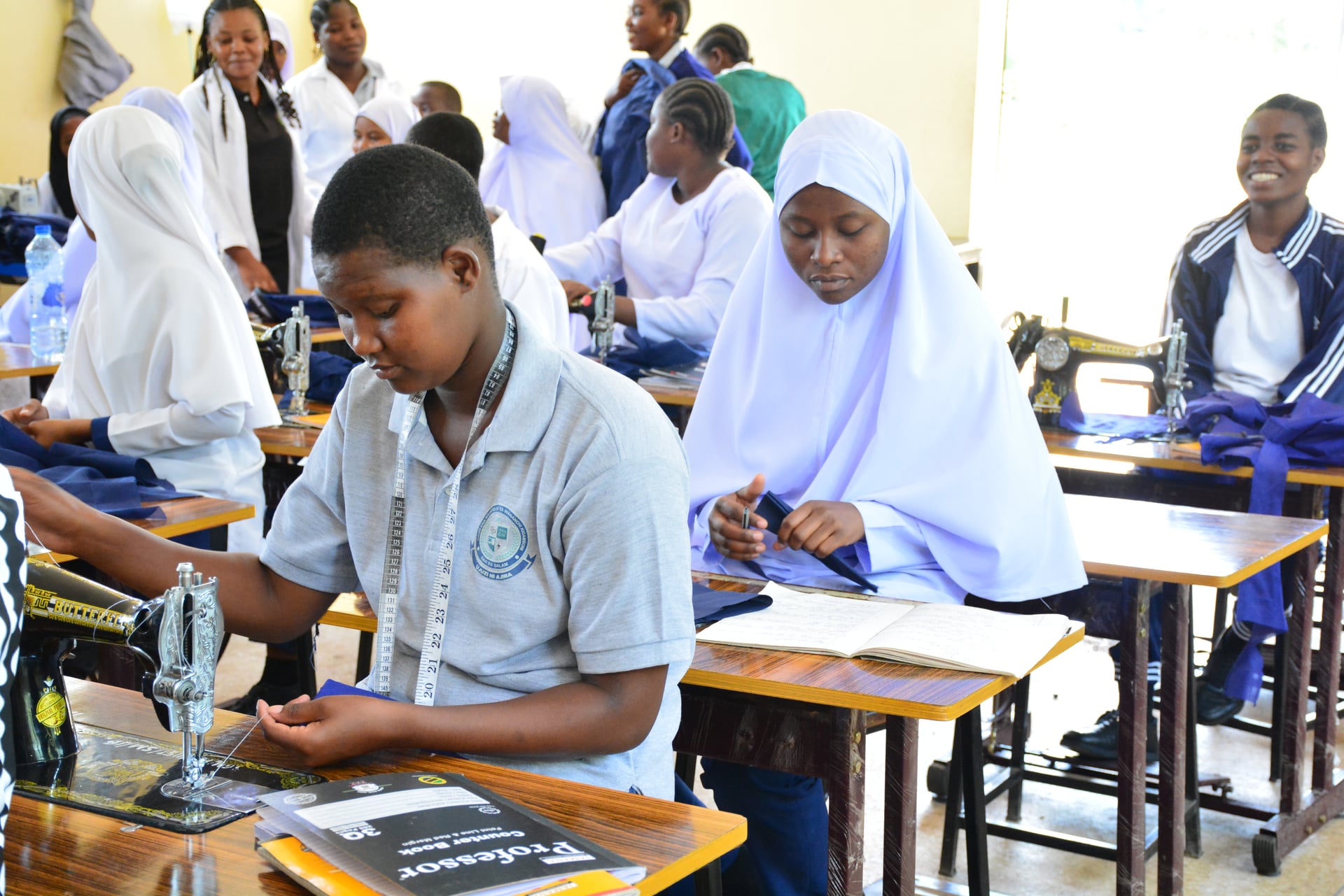 EHM students at an FDC tailoring classroom: a student in a gray polo measuring fabric at a Butterfly treadle sewing machine, with classmates in hijabs working at desks behind him