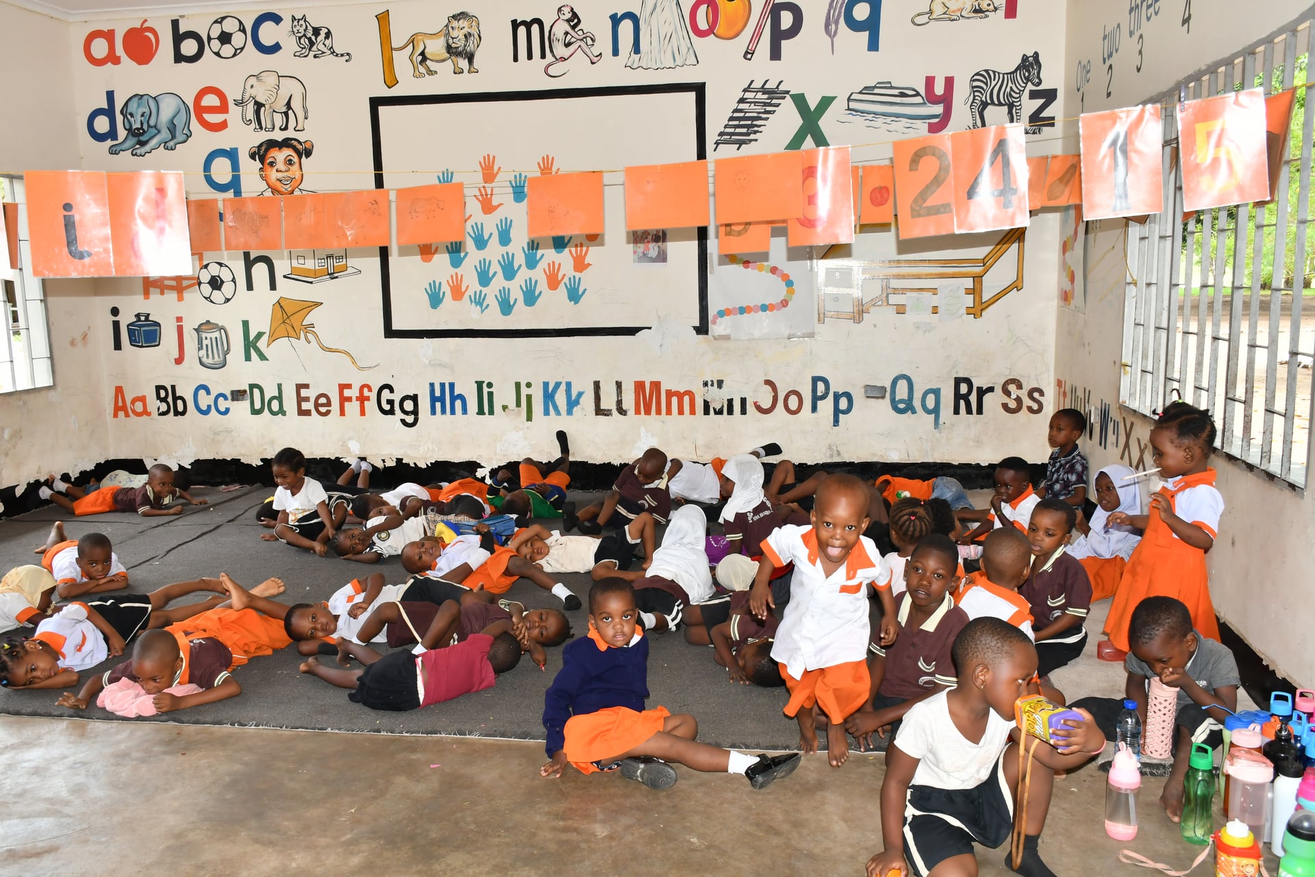 Dozens of young children rest and play in a daycare classroom decorated with hand-painted alphabet murals and colorful letter cards at an FDC center