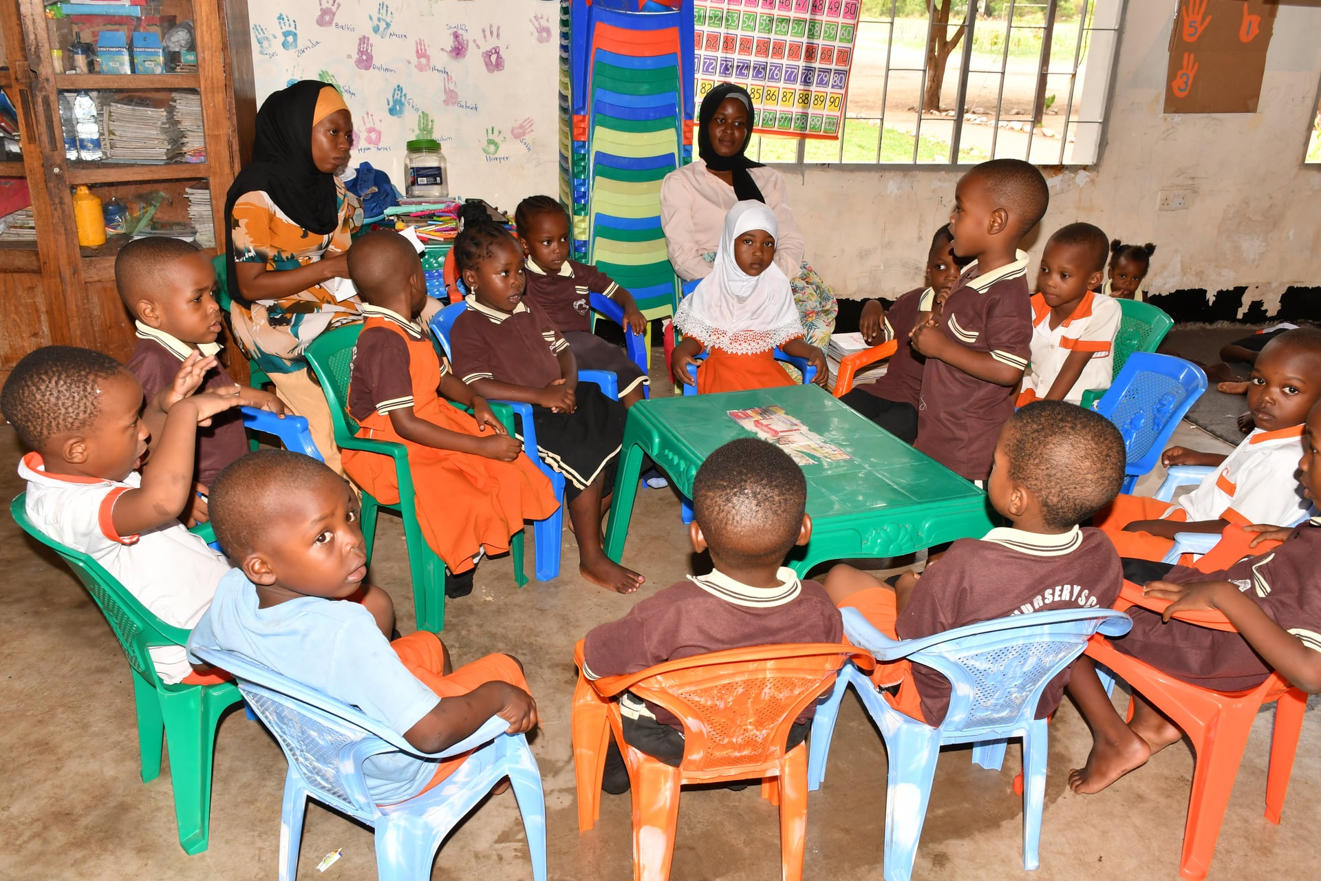 Two caregivers guide a circle of young children through a group activity at an FDC daycare center, with colorful chairs and hand-painted wall charts behind them